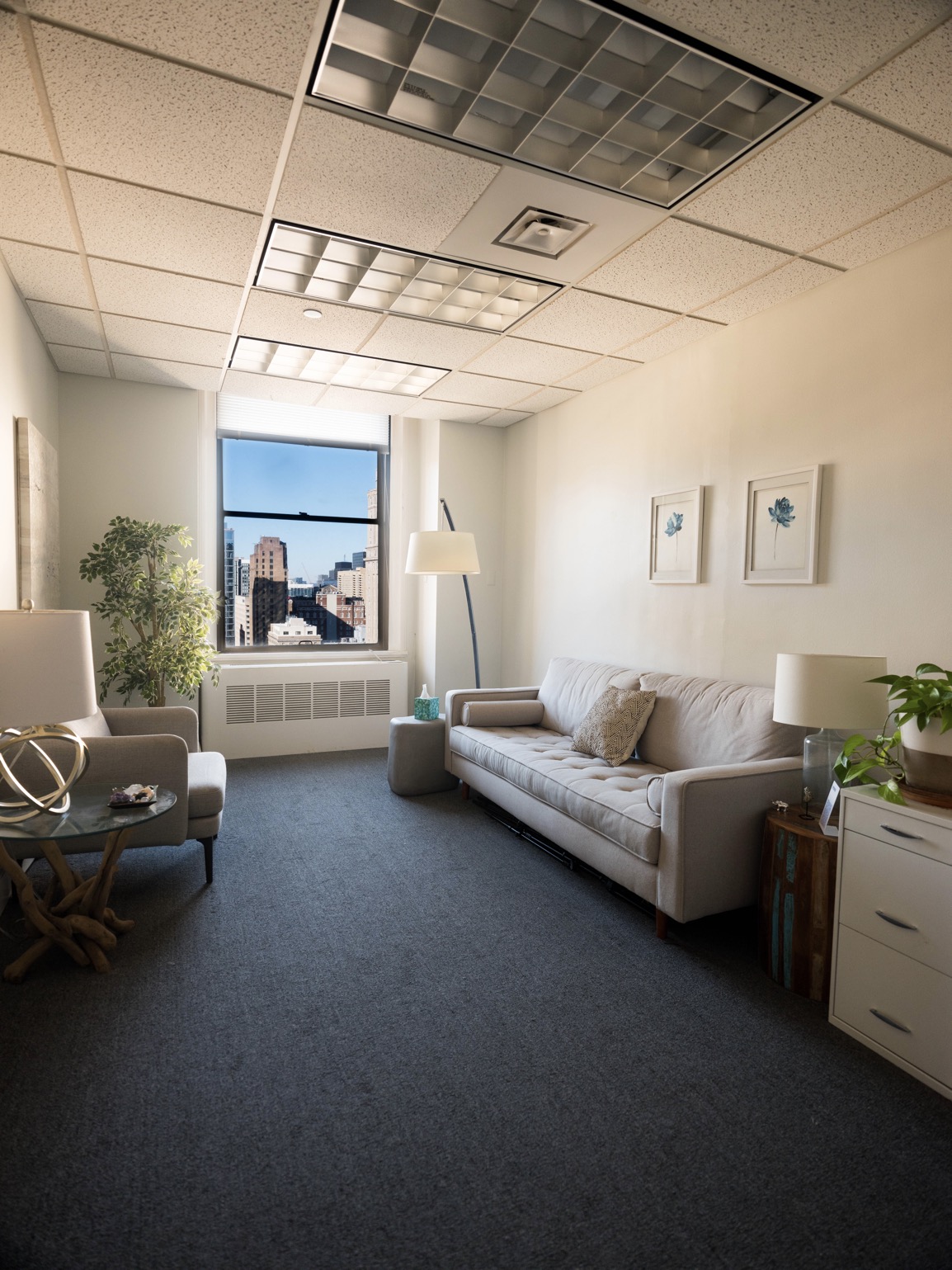 A cozy office with a large window providing a view of city buildings. The room features a comfortable sofa, a side table with a lamp, a potted plant, and framed art on the walls. There's natural light creating a warm and inviting atmosphere.