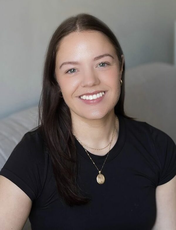 The photo shows Jordyn sitting on a light gray couch. She has long, straight brown hair and is smiling wearing a black short-sleeve top and black pants. She has a gold necklace with a round pendant, a gold bracelet, and a ring on her right hand. In the background, there is a white lamp and some green plants on a table. The setting is a cozy, well-lit room.