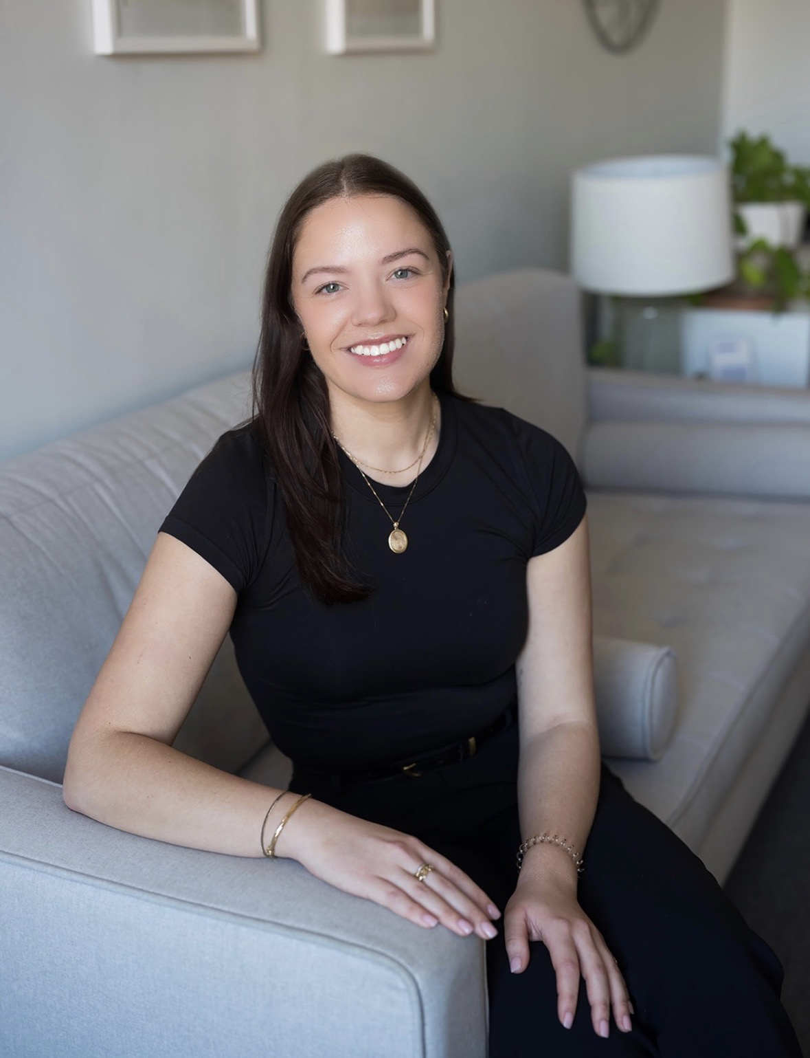 The photo shows Jordyn sitting on a light gray couch. She has long, straight brown hair and is smiling wearing a black short-sleeve top and black pants. She has a gold necklace with a round pendant, a gold bracelet, and a ring on her right hand. In the background, there is a white lamp and some green plants on a table. The setting is a cozy, well-lit room.