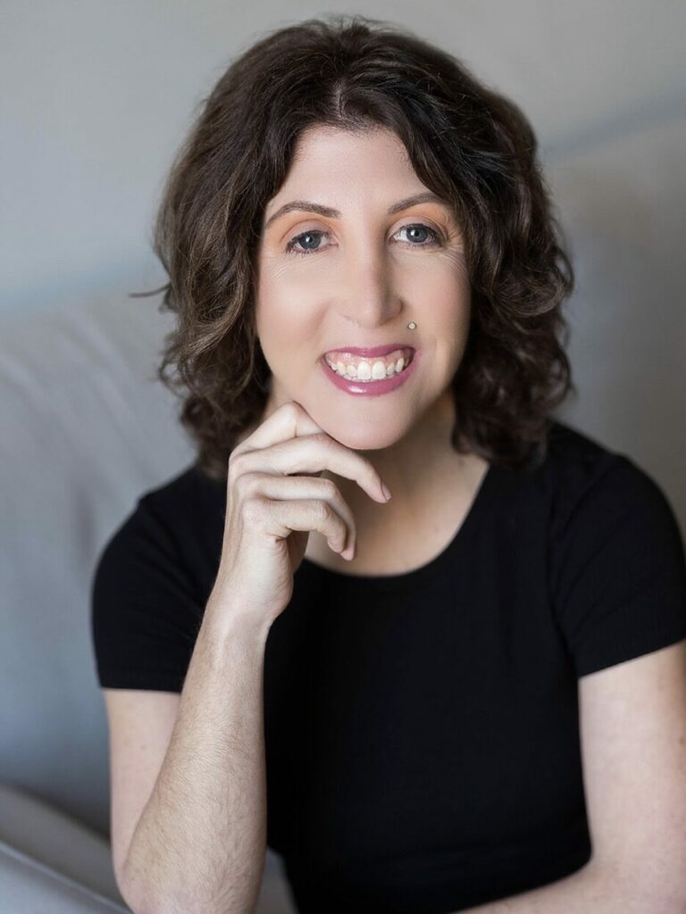 The photo shows Kimberley, with short wavy brown hair. She is smiling and has a small piercing above her lip. She is wearing a black short-sleeved top and is resting her chin on her right hand. The background is a light-grey couch.