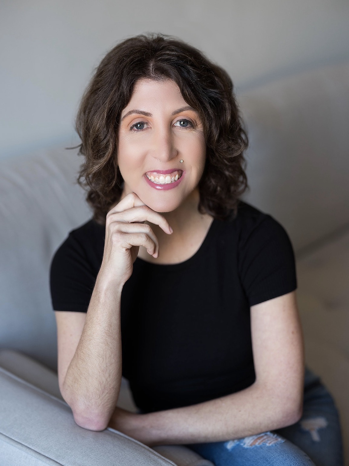 The photo shows Kimberley, with short wavy brown hair. She is smiling and has a small piercing above her lip. She is wearing a black short-sleeved top and is resting her chin on her right hand. The background is  a light-grey couch.