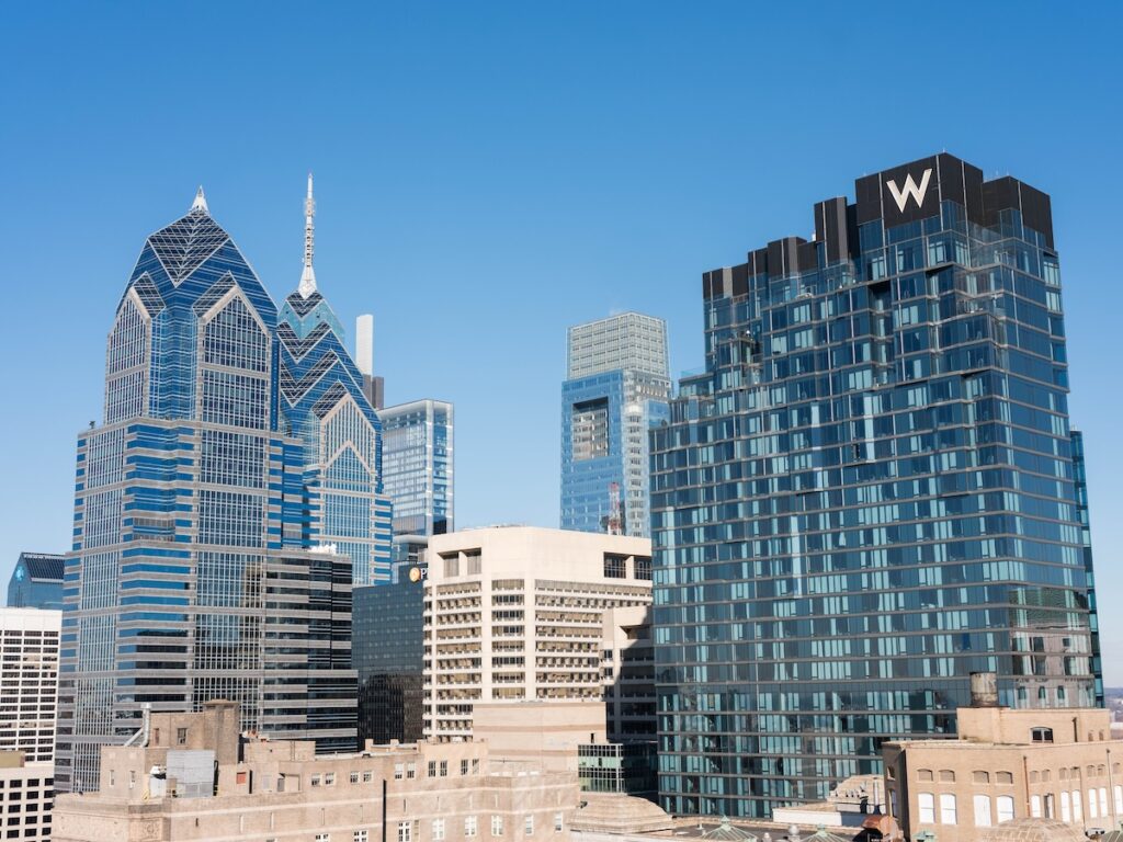 A daytime view of the Philadelphia skyline featuring modern skyscrapers, including the distinctive two-tower Liberty Place on the left and a building with a large 'W' logo on the right. The clear, blue sky enhances the striking architecture, giving a sense of home to this bustling urban landscape.