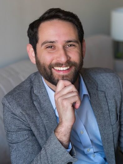 The photo shows Jacob sitting on a light-grey couch. He has short dark hair and a beard. He is smiling with his right hand resting on his chin. He is wearing a light blue dress shirt and a gray blazer. In the background, there is a white wall with two framed pictures and a table with a white lamp and some green plants. The overall setting is a cozy, well-lit room.