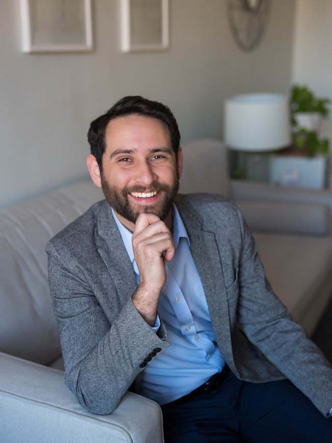 The photo shows Jacob sitting on a light-grey couch. He has short dark hair and a beard. He is smiling with his right hand resting on his chin. He is wearing a light blue dress shirt and a gray blazer. In the background, there is a white wall with two framed pictures and a table with a white lamp and some green plants. The overall setting is a cozy, well-lit room.