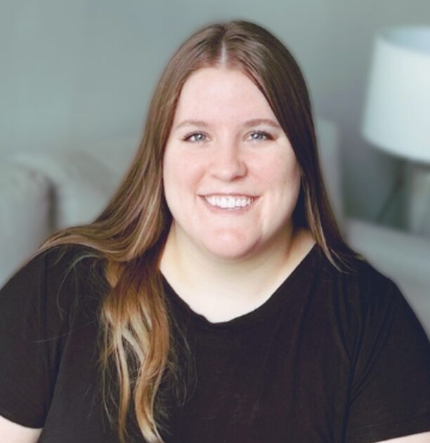 The photo shows Raegan with long, straight brown hair sitting on a light-grey couch. She is wearing a black short-sleeved shirt and is smiling. In the background, there is a white table lamp with a white shade and some green plants. The wall behind her is light gray, and there are some framed pictures.