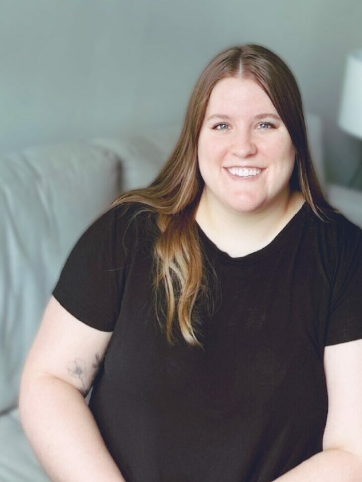 The photo shows Raegan with long, straight brown hair sitting on a light-grey couch. She is wearing a black short-sleeved shirt and is smiling. In the background, there is a white table lamp with a white shade and some green plants. The wall behind her is light gray, and there are some framed pictures.